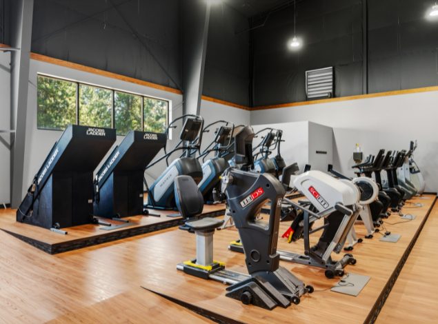 Cardio section at Peak Health & Wellness Coeur d'Alene featuring rows of treadmills and ellipticals with strength equipment and large overhead lighting in the background.