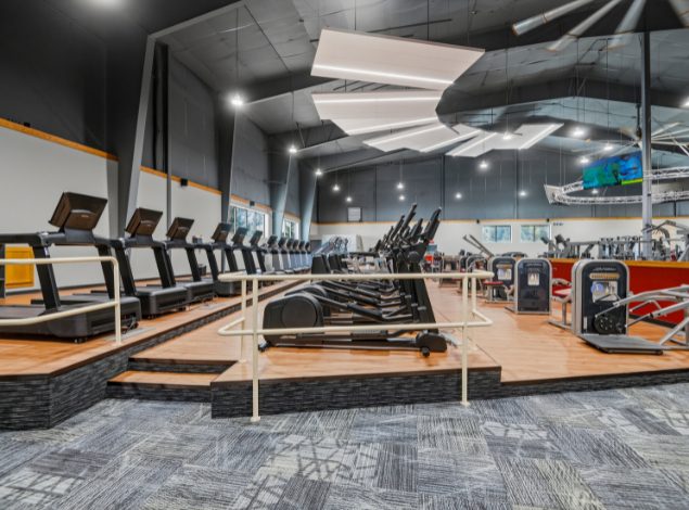Cardio section at Peak Health & Wellness Coeur d'Alene featuring rows of treadmills and ellipticals with strength equipment and large overhead lighting in the background.